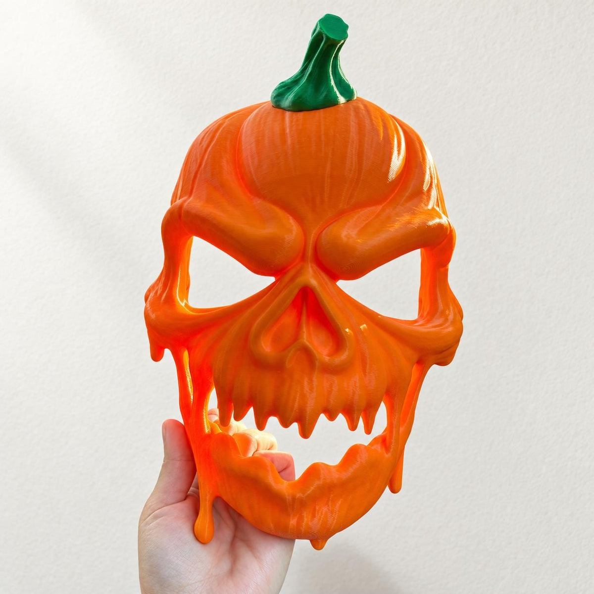 Hand holding an orange 3D printed pumpkin skull Halloween mask with a green stem